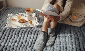 Mujer lee historias basadas en hechos reales mientras desayuna en la cama, envuelta en mantas y con zapatillas.