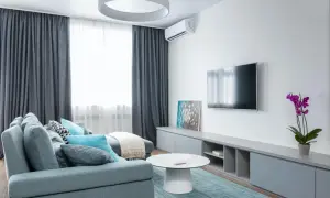 Sofá gris azulado con cojines turquesas y blancos, mesa de centro blanca, alfombra a cuadros, mueble de TV gris y orquídea morada. Un espacio que inspira una vida minimalista.