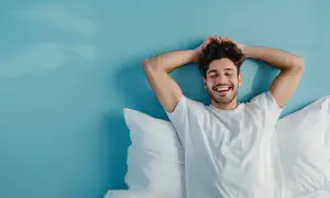 Hombre sonriente y relajado en la cama, disfrutando de levantarse a las 5 de la mañana con los brazos detrás de la cabeza.