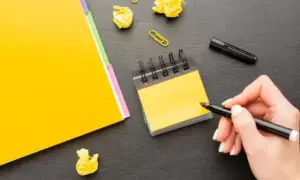 Mano escribiendo en un bloc de notas amarillo, rodeada de papeles arrugados y un marcador. Una persona proactiva planificando sus ideas.