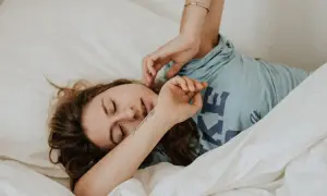 Una mujer se despierta en la cama, con los ojos cerrados y una expresión de agotamiento. Se pregunta porque está tan cansada y solo quiere dormir.