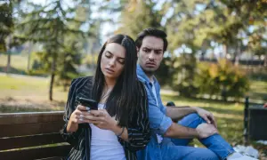 Pareja en un banco del parque, ella mira su móvil con desinterés, él la observa con recelo, reflejando que son los celos en una pareja.