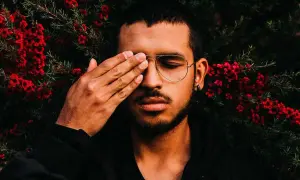 Joven con gafas y barba cubre un ojo con la mano, rodeado de flores rojas. Sus rasgos de personalidad se intuyen en su mirada introspectiva.