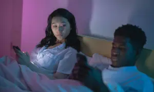 Una pareja en la cama, cada uno absorto en su teléfono. La mujer mira con recelo, quizás preguntándose qué son los celos.