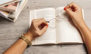 Manos con esmalte rojo sostienen unas gafas sobre un libro abierto, rodeado de más libros. Una escena que evoca la inmersión en la literatura feminista y sus historias.