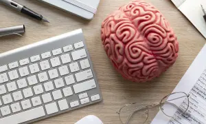 Un cerebro de juguete, teclado, bolígrafo y gafas sobre un escritorio, sugiriendo el modelo biopsicosocial.
