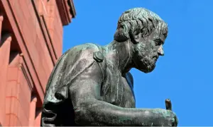 Estatua de bronce de Aristóteles, pensador cuyas frases inspiran la sabiduría. El cielo azul y un edificio de ladrillo rojo completan la escena.