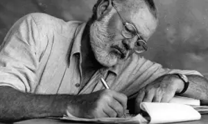 Ernest Hemingway, con gafas y barba, escribe concentrado. Sus **ernest hemingway frases** nacen de esta dedicación.