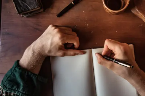 Manos escribiendo en un cuaderno abierto, buscando inspiración para frases bonitas para dedicar un libro. Una pluma, un libro antiguo y una taza de café completan la escena.