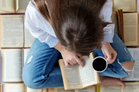 Una persona rodeada de libros que hay que leer antes de morir, con una taza de café en la mano, sumergida en la lectura.