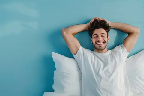 Hombre sonriente y relajado en la cama, disfrutando de levantarse a las 5 de la mañana con los brazos detrás de la cabeza.