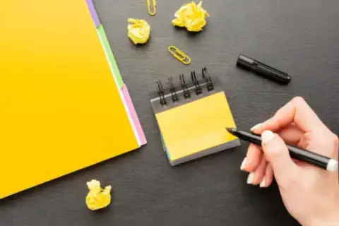 Mano escribiendo en un bloc de notas amarillo, rodeada de papeles arrugados y un marcador. Una persona proactiva planificando sus ideas.