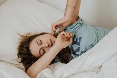Una mujer se despierta en la cama, con los ojos cerrados y una expresión de agotamiento. Se pregunta porque está tan cansada y solo quiere dormir.