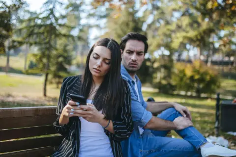 Pareja en un banco del parque, ella mira su móvil con desinterés, él la observa con recelo, reflejando que son los celos en una pareja.