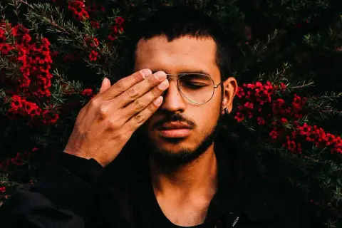 Joven con gafas y barba cubre un ojo con la mano, rodeado de flores rojas. Sus rasgos de personalidad se intuyen en su mirada introspectiva.