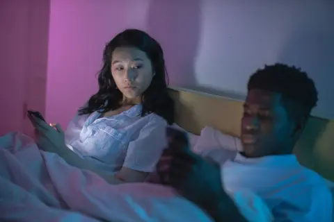Una pareja en la cama, cada uno absorto en su teléfono. La mujer mira con recelo, quizás preguntándose qué son los celos.