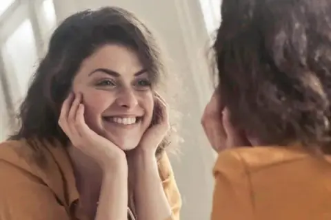 Una mujer sonríe a su reflejo, preguntándose "quien soy" mientras se mira en el espejo.