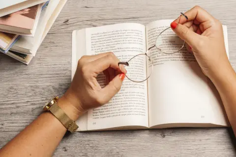 Manos con esmalte rojo sostienen unas gafas sobre un libro abierto, rodeado de más libros. Una escena que evoca la inmersión en la literatura feminista y sus historias.