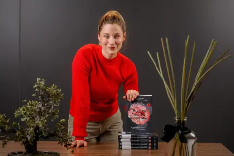 Mujer sonriente con suéter rojo, sosteniendo un libro sobre el cuerpo y la digestión, promoviendo el body positive.