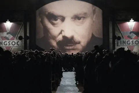 Multitud ante pantalla gigante con el rostro de un hombre y el logo de INGSOC. Un eco de las **1984 frases** que controlan sus mentes.