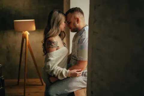 Una pareja se abraza tiernamente, sus frentes juntas. Ella tiene un tatuaje de flor en el hombro. Esto es hacer el amor de verdad: conexión y ternura.