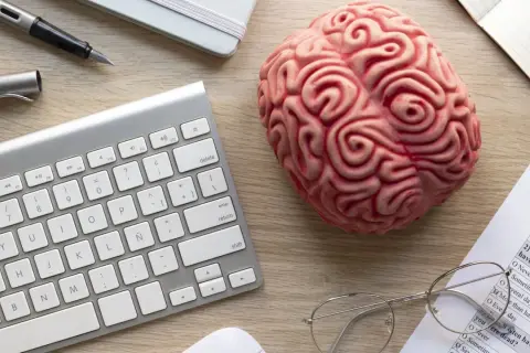 Un cerebro de juguete, teclado, bolígrafo y gafas sobre un escritorio, sugiriendo el modelo biopsicosocial.