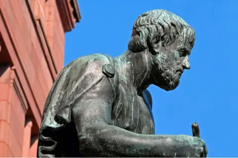 Estatua de bronce de Aristóteles, pensador cuyas frases inspiran la sabiduría. El cielo azul y un edificio de ladrillo rojo completan la escena.