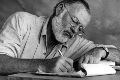Ernest Hemingway, con gafas y barba, escribe concentrado. Sus **ernest hemingway frases** nacen de esta dedicación.