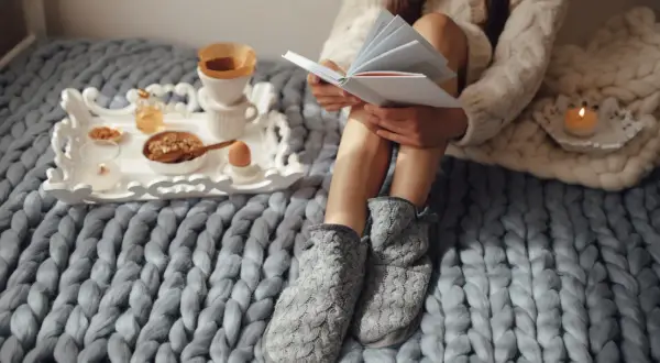 Mujer lee historias basadas en hechos reales mientras desayuna en la cama, envuelta en mantas y con zapatillas.