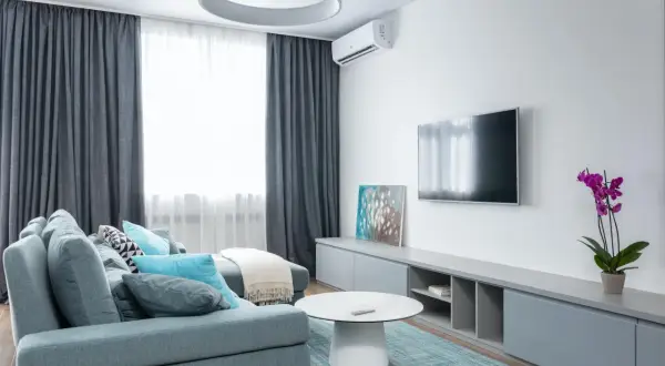 Sofá gris azulado con cojines turquesas y blancos, mesa de centro blanca, alfombra a cuadros, mueble de TV gris y orquídea morada. Un espacio que inspira una vida minimalista.