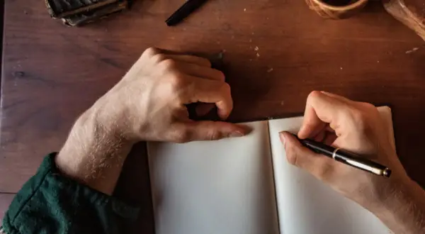 Manos escribiendo en un cuaderno abierto, buscando inspiración para frases bonitas para dedicar un libro. Una pluma, un libro antiguo y una taza de café completan la escena.
