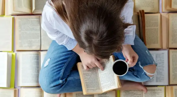 Una persona rodeada de libros que hay que leer antes de morir, con una taza de café en la mano, sumergida en la lectura.