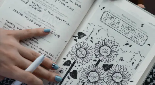 Un diario abierto muestra dibujos de girasoles y la frase "PLANT A GARDEN FULL OF SELF LOVE, GIRL!". Es un ejemplo de que es journaling, una práctica para la autoexpresión.