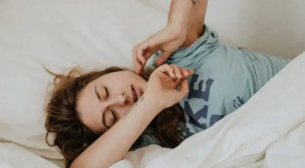 Una mujer se despierta en la cama, con los ojos cerrados y una expresión de agotamiento. Se pregunta porque está tan cansada y solo quiere dormir.