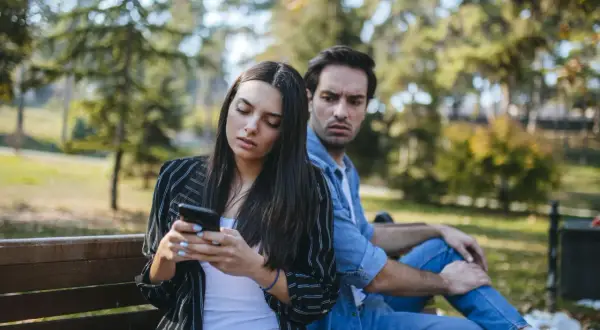 Pareja en un banco del parque, ella mira su móvil con desinterés, él la observa con recelo, reflejando que son los celos en una pareja.