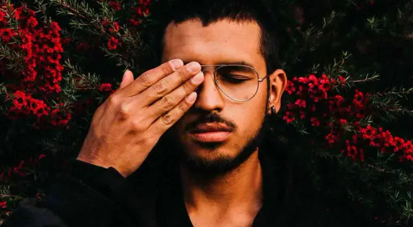 Joven con gafas y barba cubre un ojo con la mano, rodeado de flores rojas. Sus rasgos de personalidad se intuyen en su mirada introspectiva.
