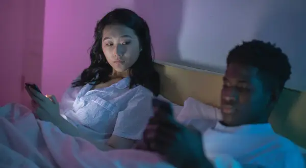 Una pareja en la cama, cada uno absorto en su teléfono. La mujer mira con recelo, quizás preguntándose qué son los celos.