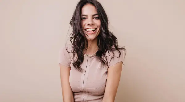 Mujer sonriendo con cabello oscuro y ondulado, luciendo un vestido beige. Su expresión irradia confianza, un ejemplo del "pretty privilege".