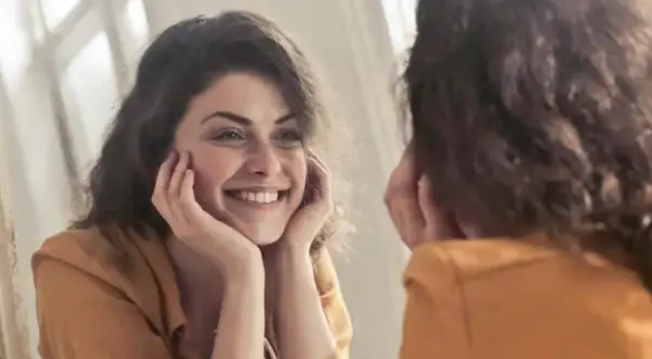 Una mujer sonríe a su reflejo, preguntándose "quien soy" mientras se mira en el espejo.