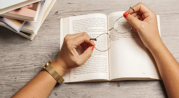 Manos con esmalte rojo sostienen unas gafas sobre un libro abierto, rodeado de más libros. Una escena que evoca la inmersión en la literatura feminista y sus historias.