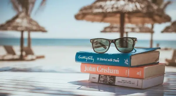Gafas de sol sobre una pila de libros para leer en inglés, con la playa y el mar de fondo.