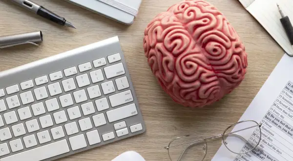 Un cerebro de juguete, teclado, bolígrafo y gafas sobre un escritorio, sugiriendo el modelo biopsicosocial.