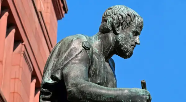 Estatua de bronce de Aristóteles, pensador cuyas frases inspiran la sabiduría. El cielo azul y un edificio de ladrillo rojo completan la escena.
