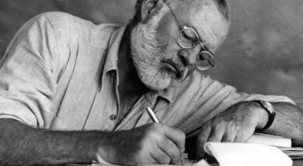 Ernest Hemingway, con gafas y barba, escribe concentrado. Sus **ernest hemingway frases** nacen de esta dedicación.