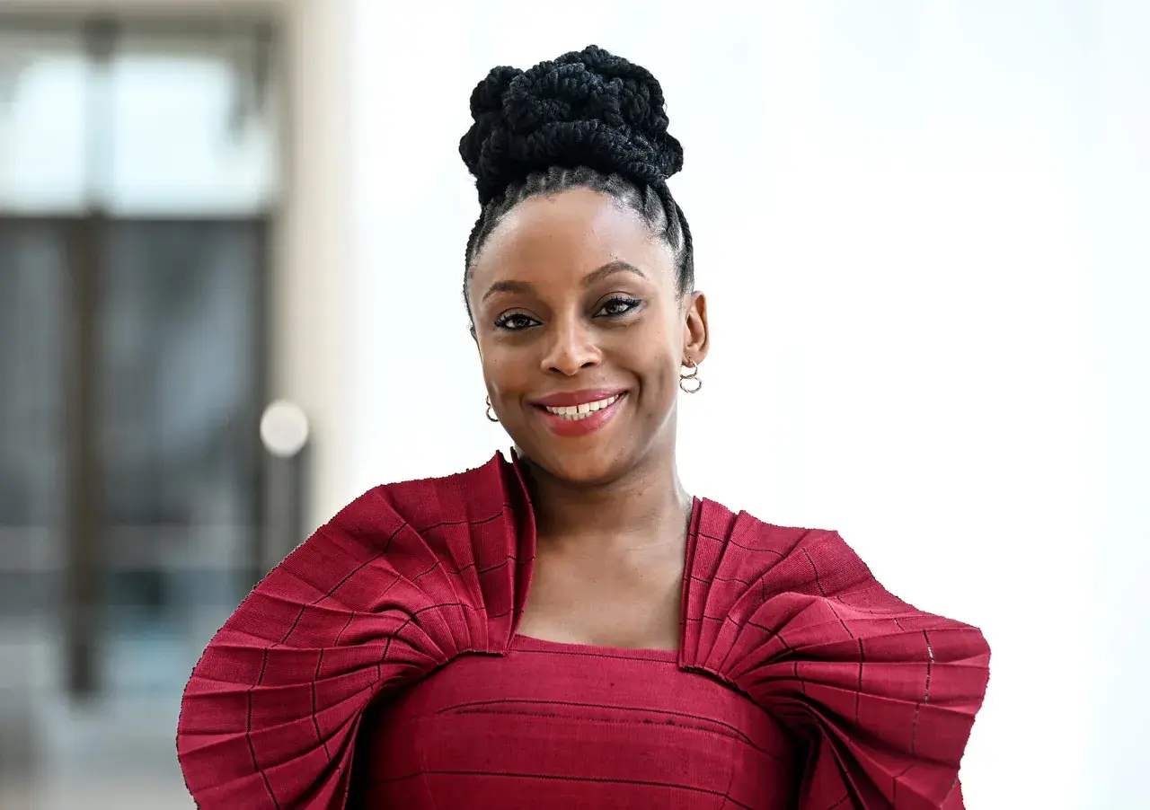 Chimamanda Ngozi Adichie, una de las escritoras famosas más influyentes, sonríe con un peinado recogido y un vestido rojo.