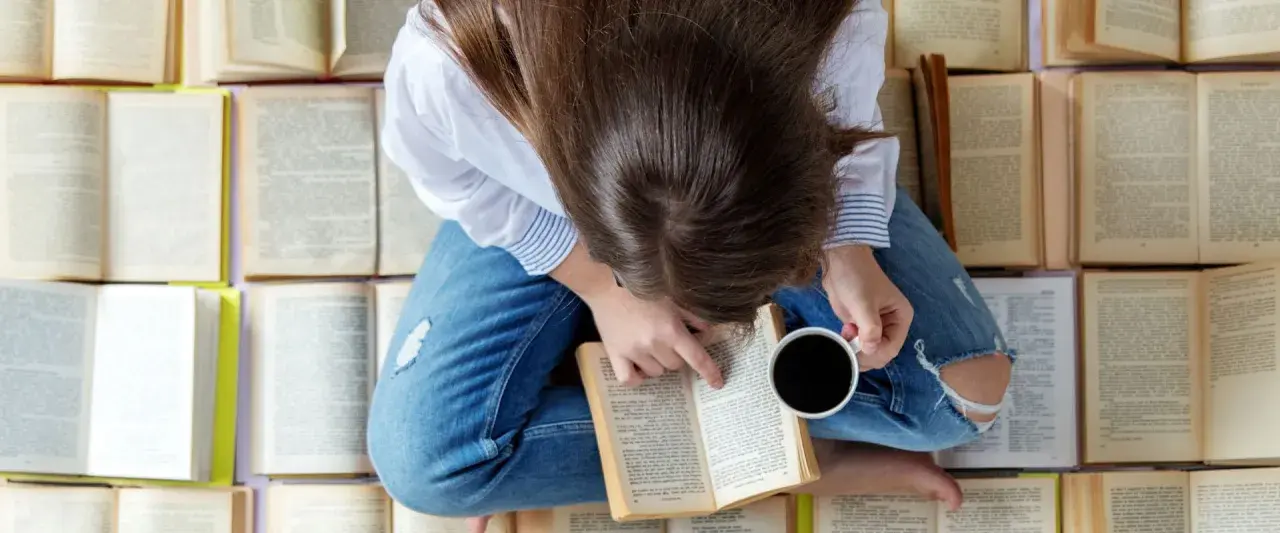 Una persona rodeada de libros que hay que leer antes de morir, con una taza de café en la mano, sumergida en la lectura.