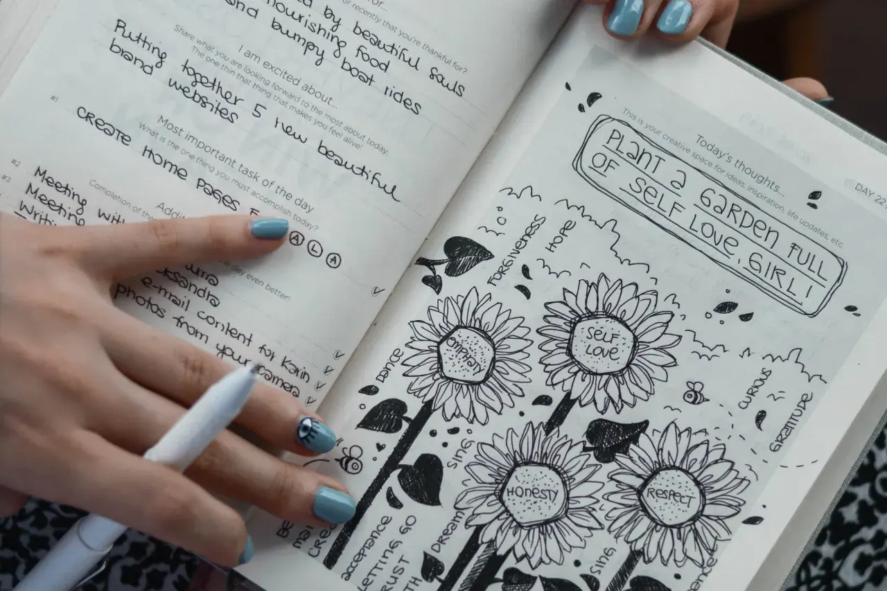 Un diario abierto muestra dibujos de girasoles y la frase "PLANT A GARDEN FULL OF SELF LOVE, GIRL!". Es un ejemplo de que es journaling, una práctica para la autoexpresión.
