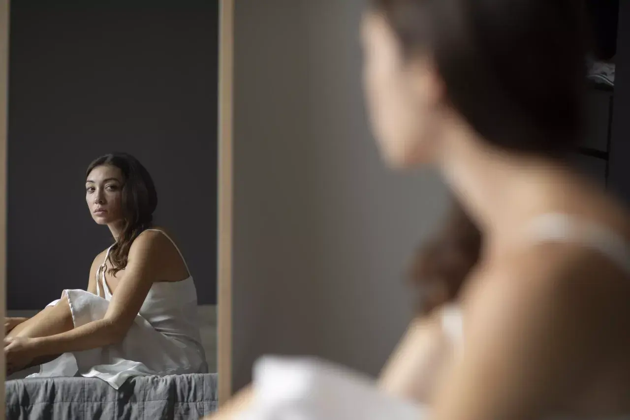 Mujer sentada en la cama, mirando al espejo con expresión de baja autoestima.