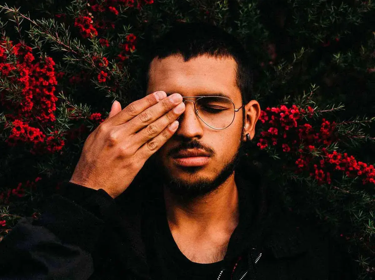 Joven con gafas y barba cubre un ojo con la mano, rodeado de flores rojas. Sus rasgos de personalidad se intuyen en su mirada introspectiva.