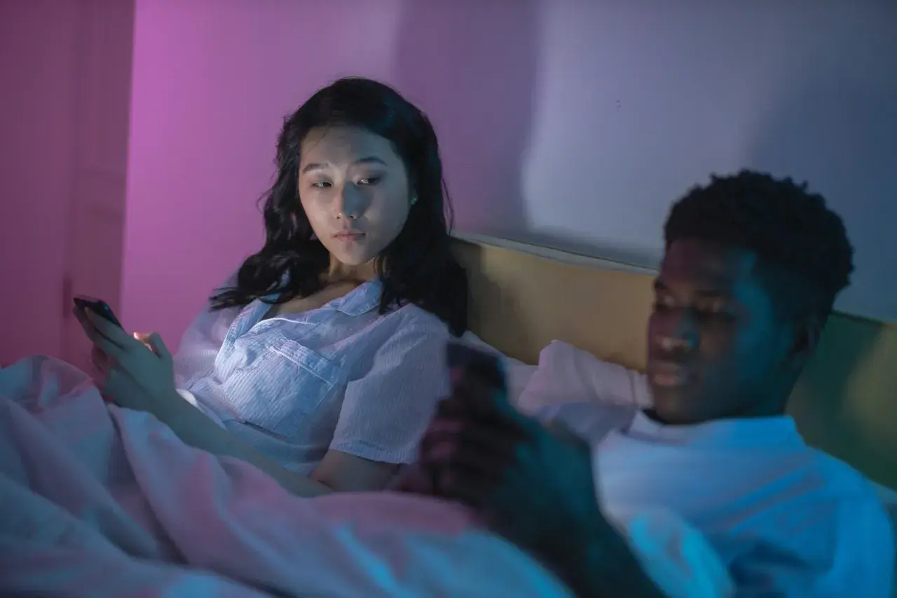Una pareja en la cama, cada uno absorto en su teléfono. La mujer mira con recelo, quizás preguntándose qué son los celos.